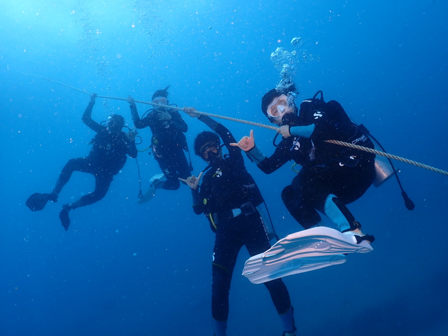 ハワイのクリアな水中へロープをつたい潜降し家族4人揃ってアロハのシャカサインを決める体験ダイバー